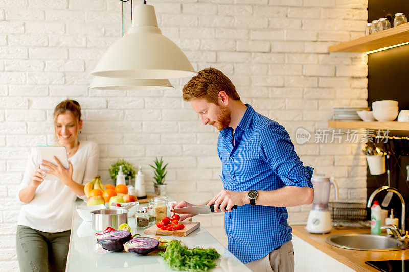 年轻夫妇有乐趣在现代厨房室内，同时准备蔬菜食物的午餐