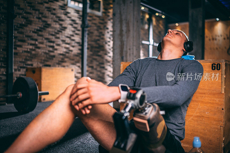 年轻的残疾运动员在健身房的地板上休息和放松，背靠在木箱上，用耳机听音乐