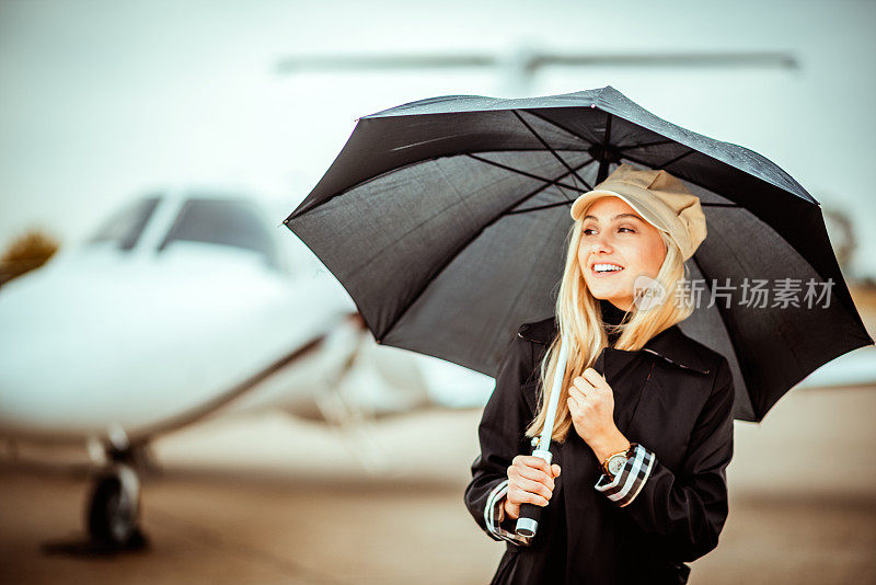 年轻的金发女模特站在机场跑道上，在一架私人飞机前，拿着一把黑色的伞