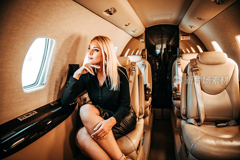 富有的金发女人在乘坐私人飞机旅行时，透过窗户看着并思考着
