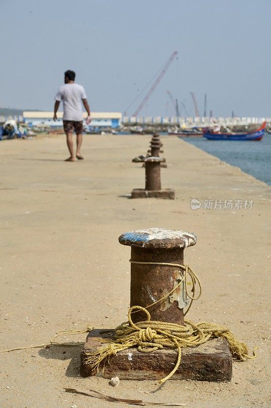 印度南部喀拉拉邦科瓦拉姆，一名印度男子走在印度渔港海港的海滨码头上，用金属柱子将系泊的渔船系在上面，系泊的船只上有起重机和远处的海鲜市场，短裤和t恤