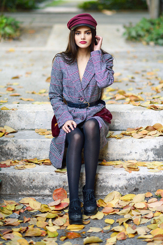 年轻漂亮的女子穿着冬衣，戴着帽子，坐在满是秋叶的台阶上