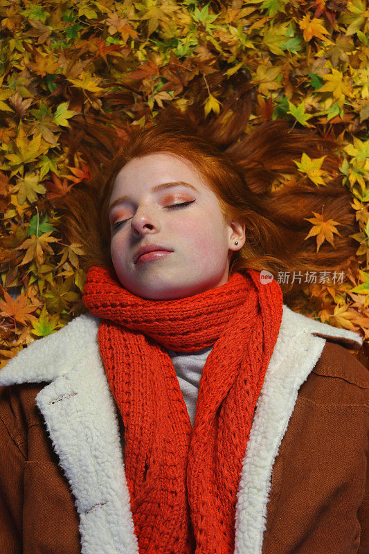 美丽的年轻漂亮的英国女孩形象与明亮的姜红色头发，长长的头发躺在黄橙色秋天枫叶外面秋天的颜色在花园在寒冷的秋天的一天，棕褐色的人造毛皮麂皮牛仔夹克，手编织橙色围巾