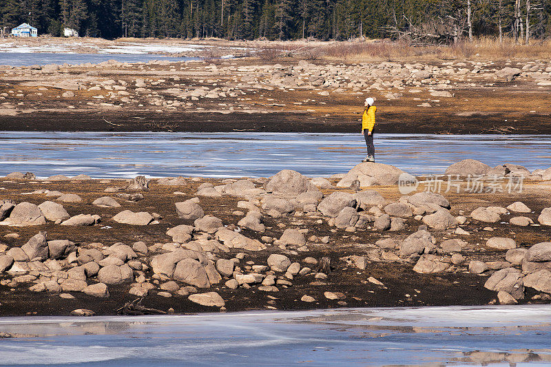 回归自然。穿着黄色夹克的独自旅行者靠近一个冰冻的山湖。一个快乐的年轻女性游客在自然。阳光明媚的户外享受。