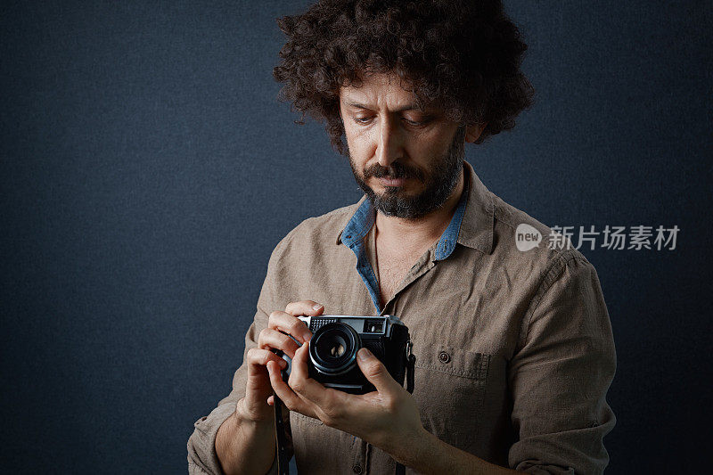 一名留着卷发的大胡子男子手持老式相机，在拍摄前调整相机，背景为深灰色