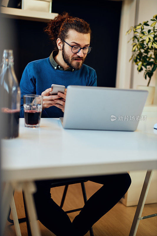 年轻帅气的大胡子嬉皮士坐在他的办公室里，喝着咖啡，用着智能手机。