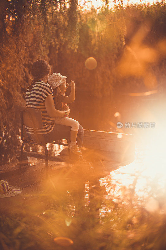快乐的幼童坐在她母亲的腿上，在湖边欣赏日落