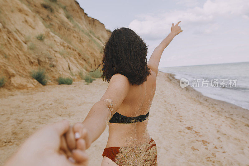 跟我来。女人牵着她的男人，在海滩上牵手。在暑假或蜜月里相爱的情侣。年轻女子与棕褐色的身体握着男人的手