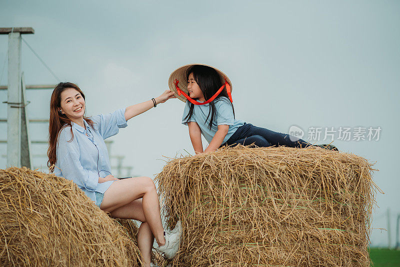 亚洲华人女儿微笑着坐在草垛上，帮助母亲戴上传统的藤帽