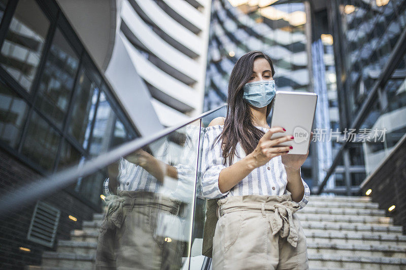 美丽的女人站在城市街道上使用数字平板电脑，脸上戴着防护面具。病毒大流行与污染概念。