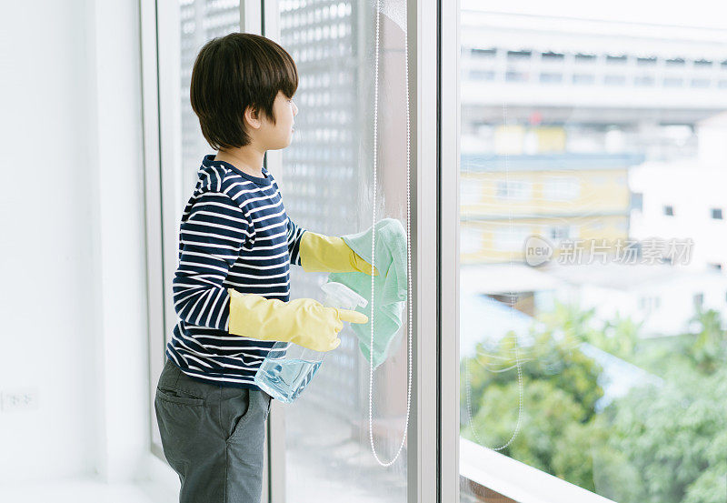 侧面的亚洲小男孩戴着橡胶手套做房子清洁在客厅清洗窗户。人、小孩、家务和家务观念。
