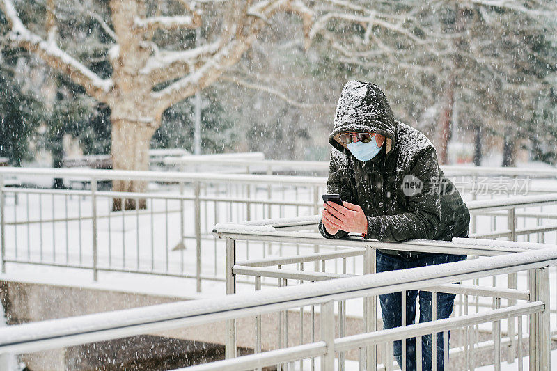 年轻人戴着防护口罩和太阳镜在下雪天使用手机