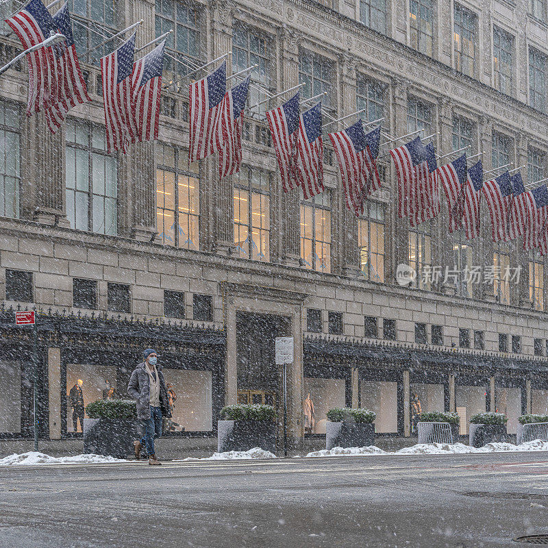 一名戴着防护口罩的年轻人正在恶劣的天气中穿过曼哈顿的第五大道，当时正值冬季暴风雪和大雪。