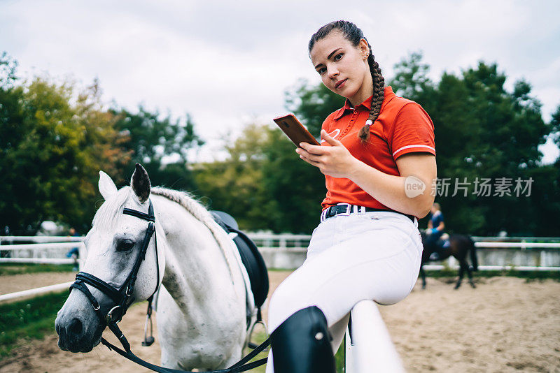 女人坐在围栏附近的马在跑道上使用智能手机