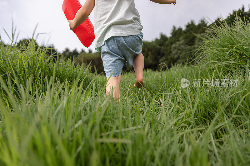 快乐童年理念，探索你身边的世界。一个可爱的女孩，白天光着脚在公园的草地上跑步，手里拿着一个气球。