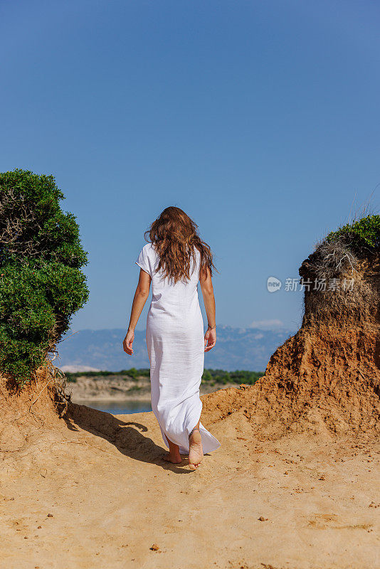 身穿夏日长裙的黑发美女后视图，正沿着海岸的旱地往上走