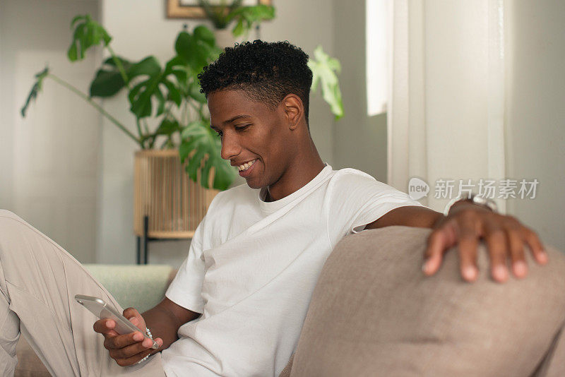年轻人，在沙发上玩智能手机和社交媒体，周末在家看视频放松一下。学生，微笑和手机用于在线研究和公寓健康的教育应用程序