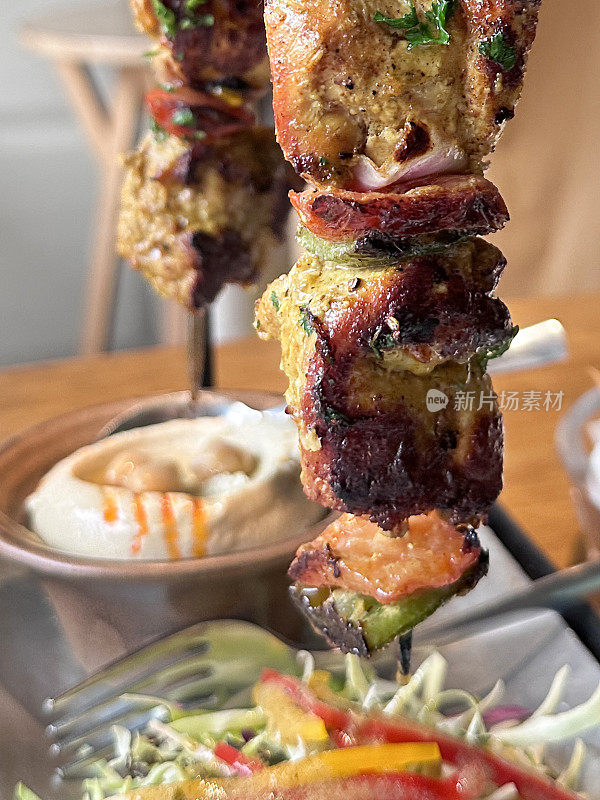 两张特写图片，悬挂在金属串架上的鸡肉串，烤焦的鸡胸肉块和覆盖着辣酱的红青椒串在一起，一碗香浓汤，餐厅设置背景，重点放在前景上