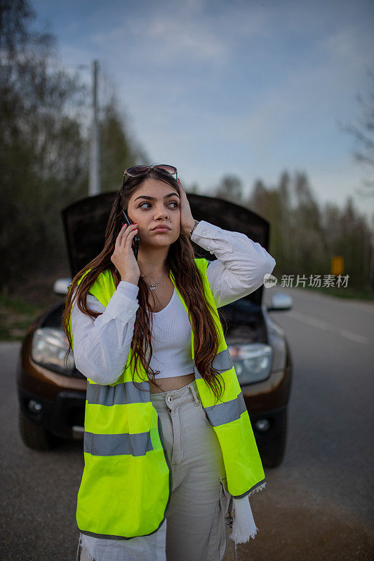 年轻的压力很大的女子站在她抛锚的汽车旁边，用手机求救