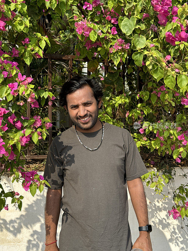 一名印度男子站在阳光下，在粉红色的九重梅花的背景前摆姿势，这些花生长在葡萄藤上，就像不寻常的热带开花花园篱笆，重点放在前景上