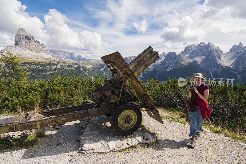 在意大利Dolomites的皮亚那山上，一位年长的妇女带着相机走过一战时期的大炮，背景是拉瓦雷多城