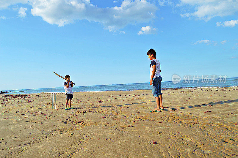 在一个阳光明媚的夏日，两个印度小孩穿着相同的t恤在海边的沙滩上打板球。