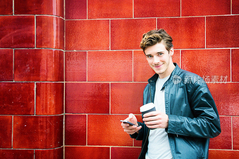 千禧一代的英国男人肖像靠在红色的墙上，拿着电话和咖啡