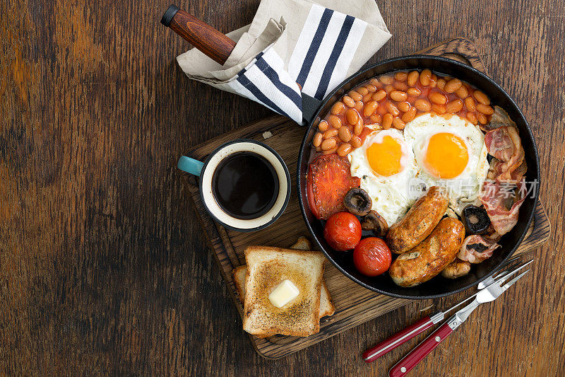 完整的英式早餐，有香肠，培根，煎蛋，豆子，吐司和一杯咖啡，放在有复印空间的木桌上