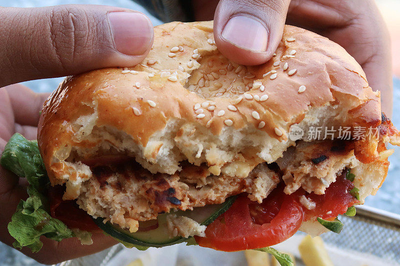 这是一名面目全非的男子手持的芝麻面包鸡肉汉堡，里面有鸡肉肉饼、洋葱圈、番茄片、黄瓜和莴苣叶，还有番茄酱