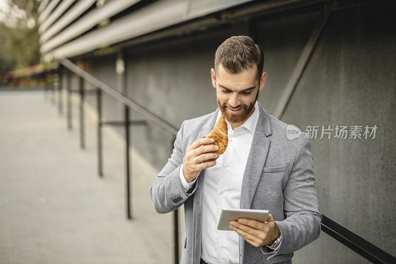 商人工作的肖像，使用数字平板电脑和吃牛角面包的户外