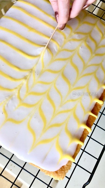 图片:一个不认识的人用鸡尾酒棒在白色方糖糖霜上画出黄色柠檬味糖霜的羽毛图案，自制的柠檬托盘烤蛋糕放在金属冷却架上，高架视野