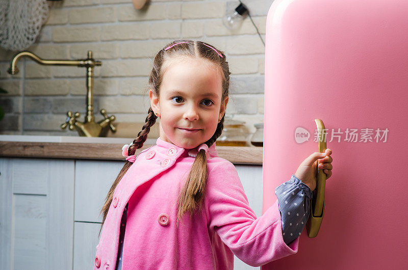 在一间复古风格的厨房里，一个穿着粉色外套的五岁小女孩打开了一台老式的粉色冰箱。