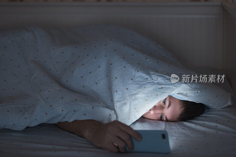 人像困倦的男子躺在床上盖着毯子用智能手机，深夜无法入睡。失眠，无手机恐惧症，睡眠障碍的概念。