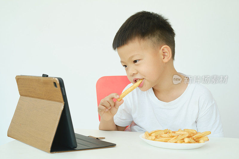 一个亚洲男孩一边吃薯条，一边看平板电脑上的视频