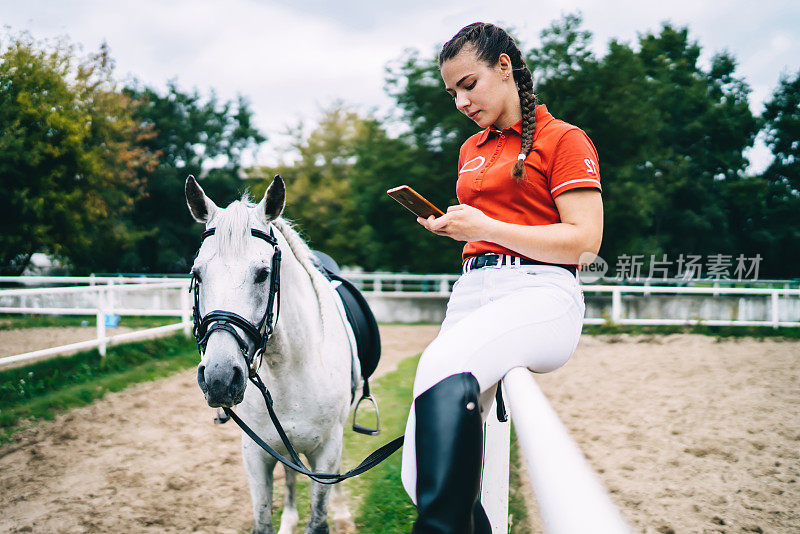 女骑师坐在马旁边的栅栏上使用智能手机