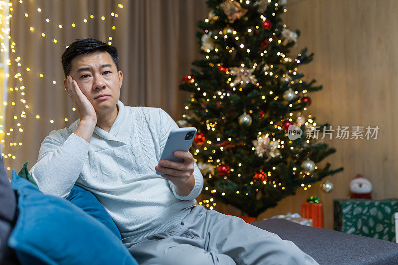 一位年轻的亚洲男子坐在家里，手里拿着手机。等待一个电话，通知