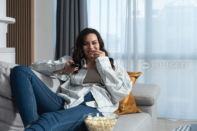 年轻的职业女性在沙发上休息，用遥控器在电视菜单上选择电视节目或电影。自由女性独自在家度过一天，周末活动概念