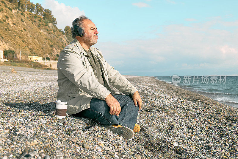 快乐的中年大胡子男子坐在冬天的海滩上，戴着耳机听着智能手机应用程序的音乐或播客。