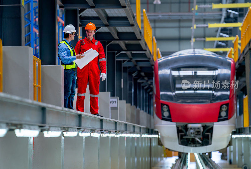专业工程师手持图纸图纸，技术工人在工厂车间电气或空轨列车的铁轨旁停留讨论。