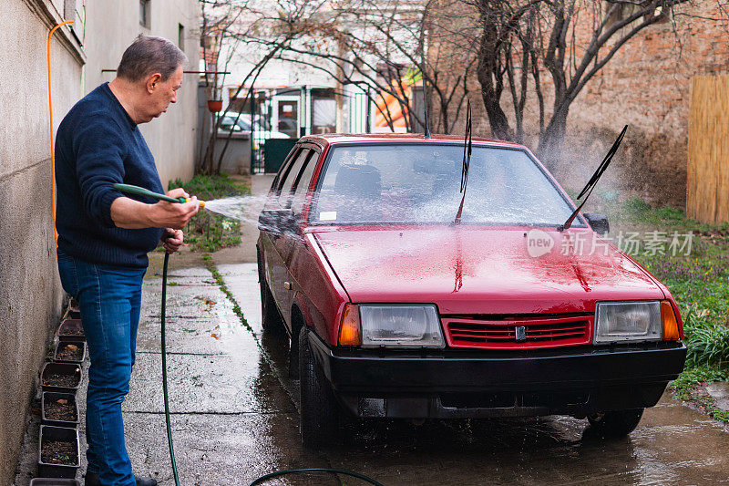 一位老人手里拿着一根软管，在他的后院洗车。