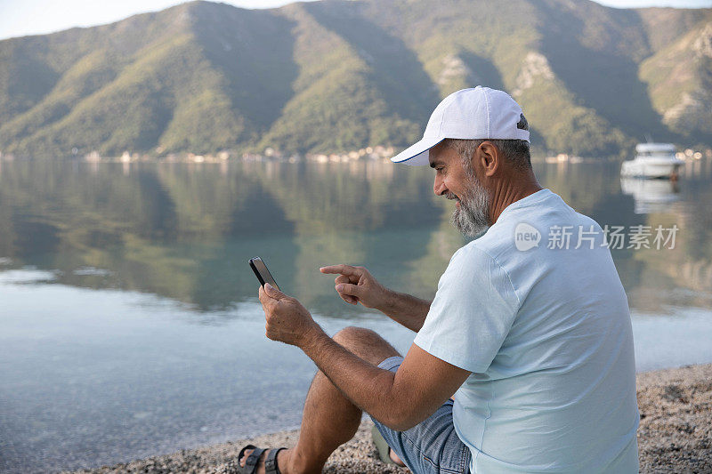 一位白发苍苍的老人坐在海边的长椅上，微笑着看着自己的智能手机。迷人的人们微笑着享受户外和大自然的美丽。老年人和现代科技股票照片…