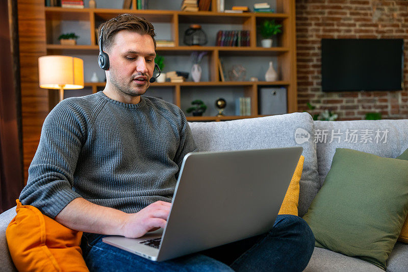 男人坐在家里客厅的沙发上，喜欢用笔记本电脑和耳机学习，看着设备屏幕，听音频，做一些笔记。在互联网概念中有在线在线教学的概念