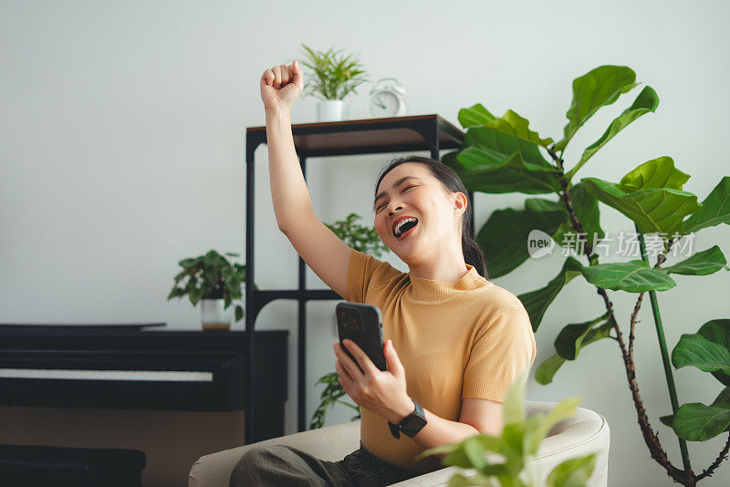 亚洲女性坐在家里的扶手椅上，用智能手机开心兴奋地做出了获胜的手势。