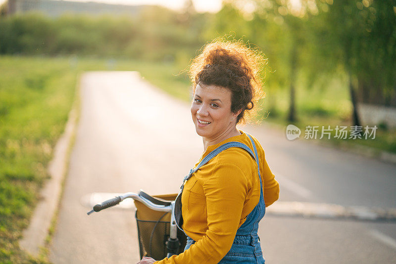 在阳光明媚的公园里，穿着黄色衬衫、卷头发的微笑女子推着自行车