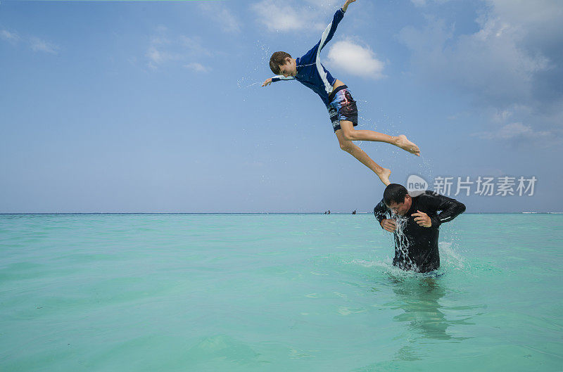 马尔代夫古里希岛海滩上，小男孩像鸟儿一样，从男人的肩膀上跃入水中，背靠蓝天，享受着碧蓝的海水