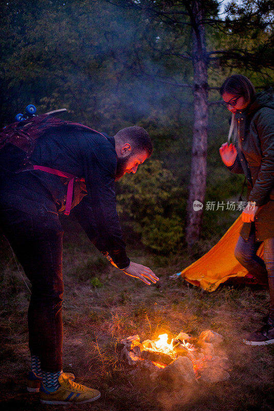 漂亮的一对，围坐在篝火旁。背景是一个帐篷，那人用一根棍子弹火。