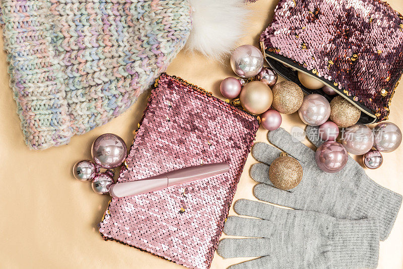 俯视图的圣诞女孩时尚构成，闪闪发光的记事本与钱包，针织帽子和手套在粉红色和灰色的金色背景