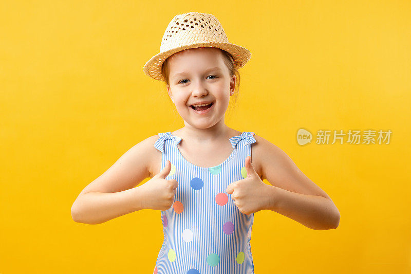快乐可爱美丽迷人的小女孩戴着草帽和泳衣显示拇指向上的双手。黄色背景上的一个孩子的特写肖像