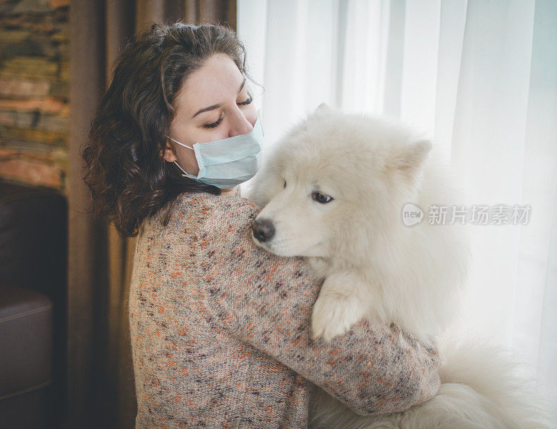 年轻女子戴着医用口罩，带着狗站在窗边