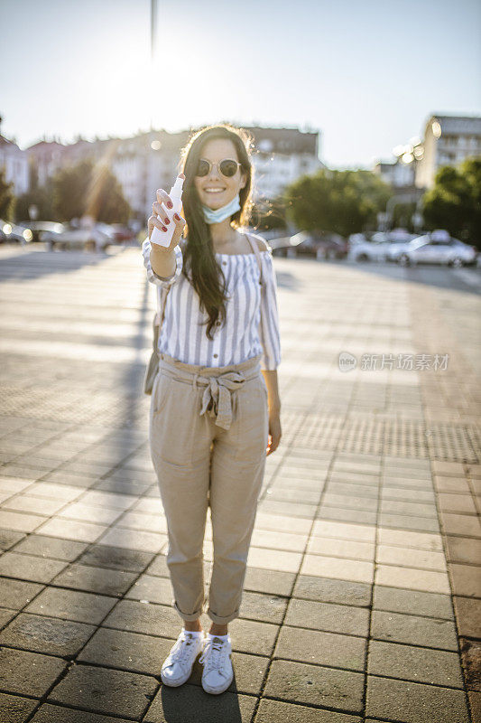 一名年轻女子戴着外科口罩，手上涂着洗手液，在封锁期间站在空荡荡的城镇广场上